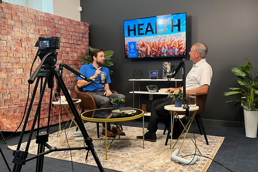 Wyatt Beckman and Dr. Robert Moser recording a podcast in a studio, seated with microphones. A camera on a tripod captures the conversation. The screen behind them reads 'Health on the Plains,' with a graphic of wheat fields. The studio is decorated with plants and a brick wall backdrop.